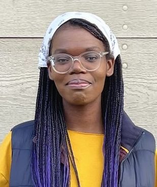 Caroline Imani Charles stands against a light wall wearing white head scarf, eyeglasses, and a blue vest over a yellow shirt