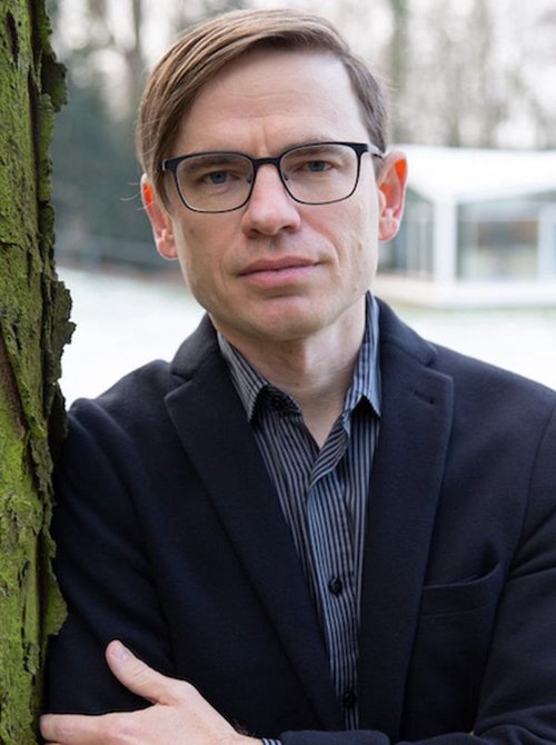 Author Jared Farmer, in striped shirt and blue jacket leans against a green-barked tree against a snowy white background