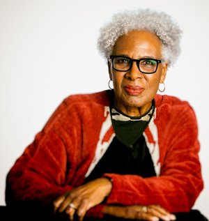 Nell Irvin Painter wears red-orange sweater and dark framed eyeglasses, arms crossed in front of her on a table