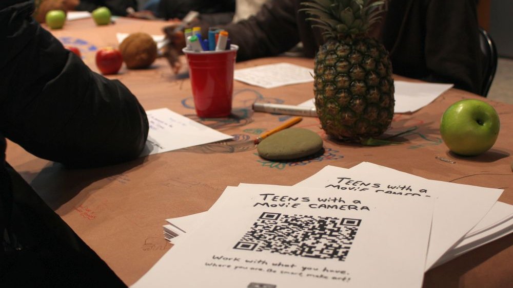 a stack of fliers that say "Teens with a Camera" lays on corner of a table where people are writing