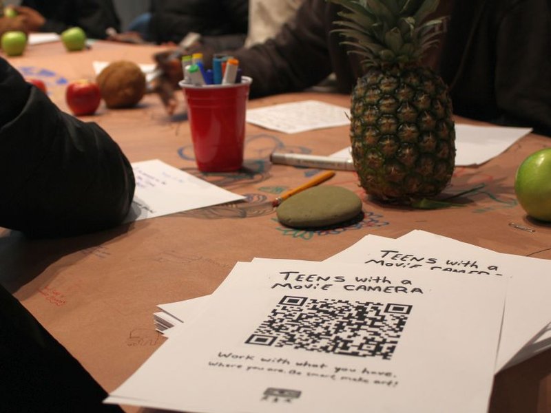 a stack of fliers that say "Teens with a Camera" lays on corner of a table where people are writing